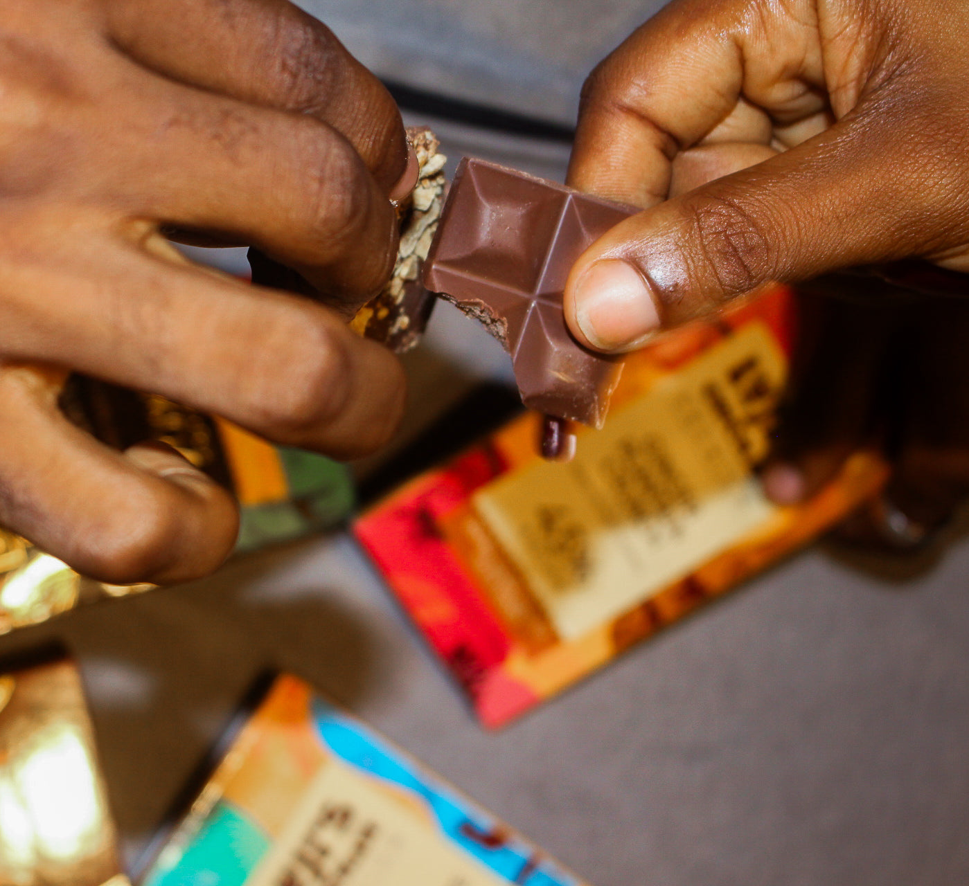 Close-up of two hands breaking a Luji’s Chocolate bar over a table filled with colorful West African–inspired chocolate packaging.