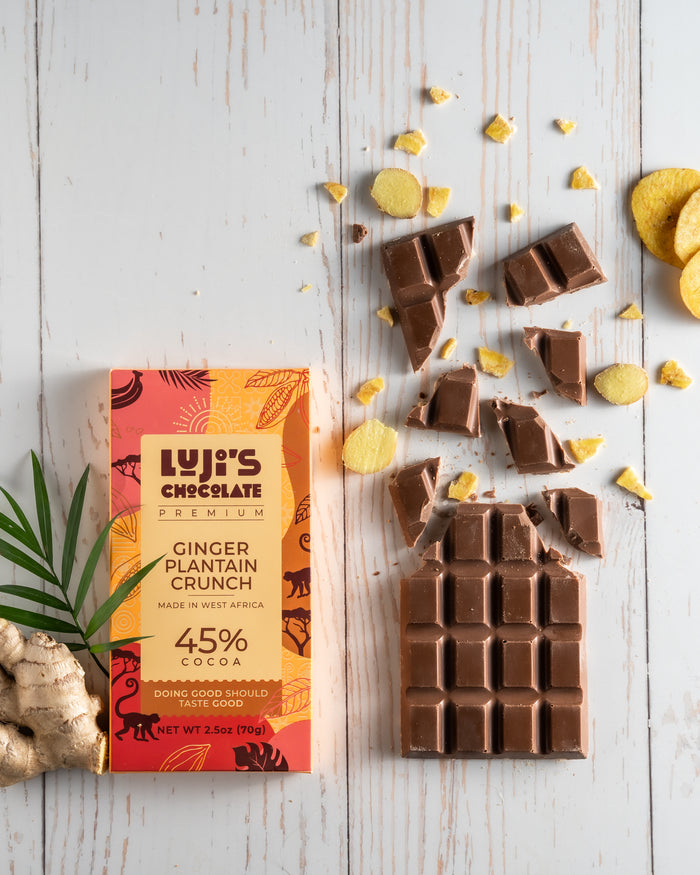 An unwrapped Luji's Ginger Plantain Crunch Chocolate bar broken into pieces alongside crispy plantain chips and fresh ginger, illustrating the bar's flavor components on a rustic white wooden background.