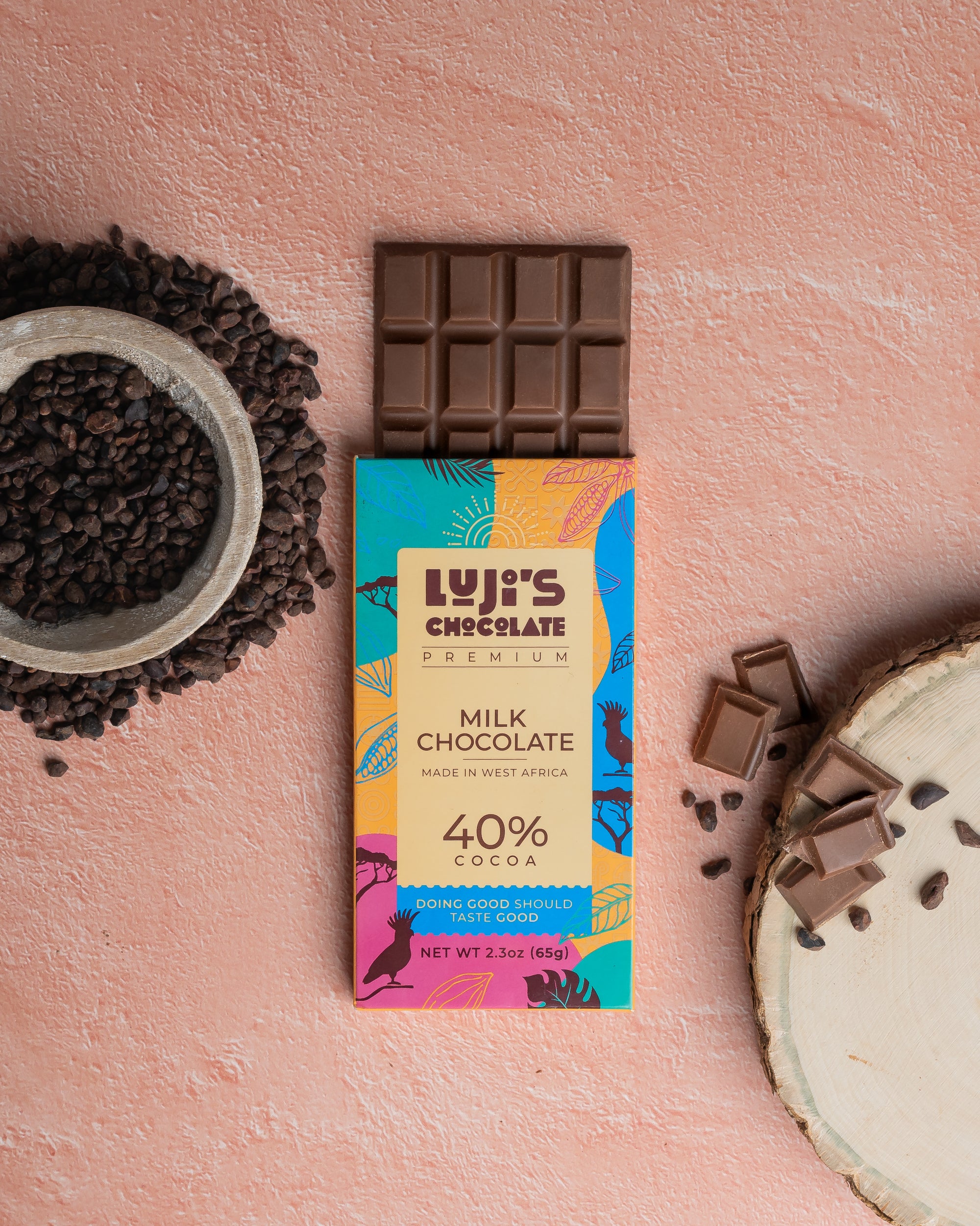 Luji's Chocolate Milk Chocolate bar opened beside a bowl of cocoa beans, with chocolate chunks on a cut tree stump, conveying the natural and pure ingredients used in the chocolate against a pink backdrop."