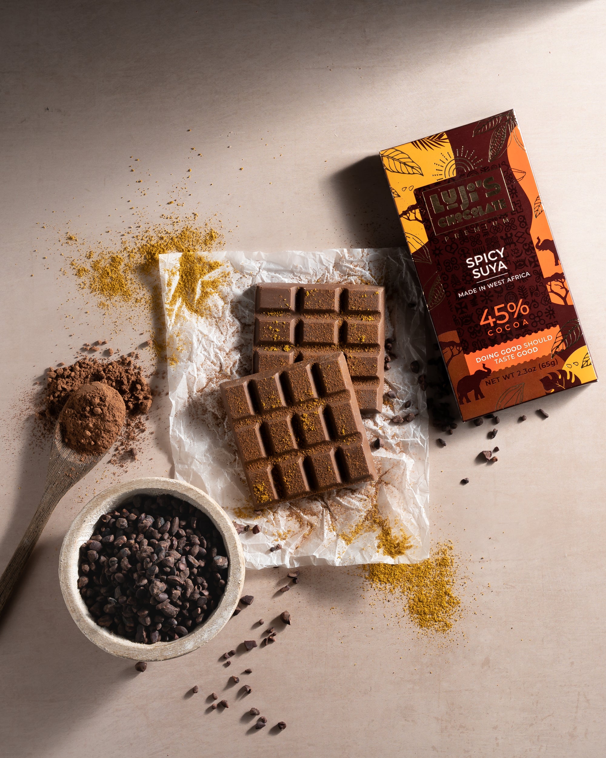 Luji's Spicy Suya Chocolate bar displayed with a spoonful of suya spices and cocoa nibs, highlighting the spicy and chocolaty elements that make up the bar's unique flavor, on a parchment paper backdrop.